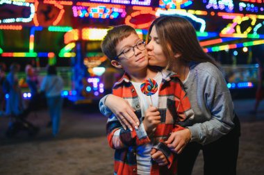Sevimli bir çocuk ve annesi renkli bir lolipop yiyorlar. Atlıkarıncanın arka planında duruyorlar.
