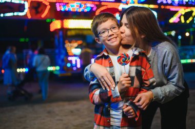Sevimli bir çocuk ve annesi renkli bir lolipop yiyorlar. Atlıkarıncanın arka planında duruyorlar.