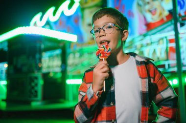 Akşamları eğlence parkında ya da sirkte renkli bir lolipop yiyen beyaz bir çocuk..