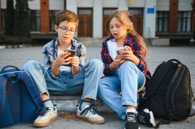 Modern okul binasının yanındaki merdivenlerde oturan iki küçük öğrenci. Akıllı telefondan interneti arayan ve arkadaşına gösteren güzel arkadaşlar.