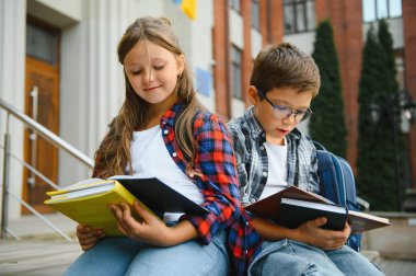 Bir grup ilkokul çocuğu kitap okuyorlar. Modern okul binası arka planında açık hava sınıfları var..