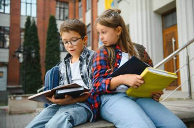 Bir grup ilkokul çocuğu kitap okuyorlar. Modern okul binası arka planında açık hava sınıfları var..