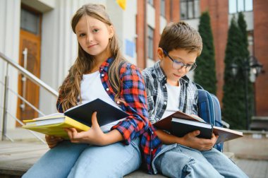 Bir grup ilkokul çocuğu kitap okuyorlar. Modern okul binası arka planında açık hava sınıfları var..