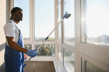 Önlüklü Afrikalı profesyonel temizlik işçisi pencereleri özel ekipmanlarla temizliyor..