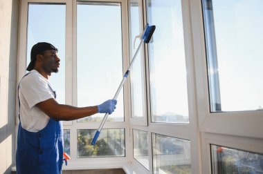 Tulumlu Afrikalı profesyonel temizlik işçisi, özel ekipmanlı bir mağazanın camlarını ve pencerelerini temizliyor..
