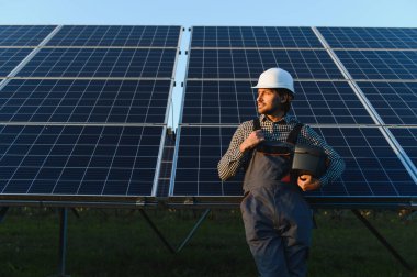Güneş panelinin arka planında duran üniformalı ve kasklı genç profesyonel bir işçinin portresi..