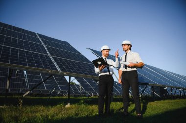Laptop kullanan ve projeyi tartışan erkek mühendisler kırsal kesimde fotovoltaik pillerin yanında duruyorlar..