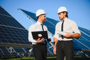 Laptop kullanan ve projeyi tartışan erkek mühendisler kırsal kesimde fotovoltaik pillerin yanında duruyorlar..