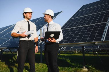 Laptop kullanan ve projeyi tartışan erkek mühendisler kırsal kesimde fotovoltaik pillerin yanında duruyorlar..