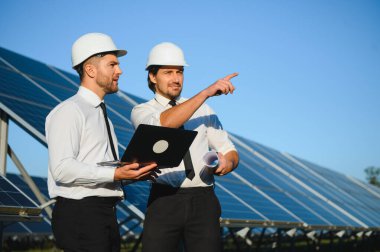 Laptop kullanan ve projeyi tartışan erkek mühendisler kırsal kesimde fotovoltaik pillerin yanında duruyorlar..