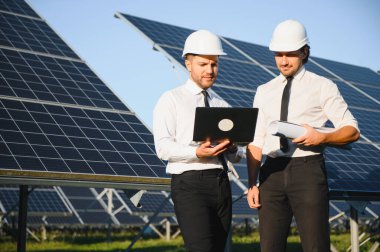 Laptop kullanan ve projeyi tartışan erkek mühendisler kırsal kesimde fotovoltaik pillerin yanında duruyorlar..