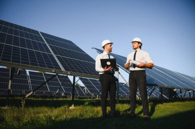 Laptop kullanan ve projeyi tartışan erkek mühendisler kırsal kesimde fotovoltaik pillerin yanında duruyorlar..