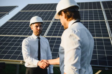 Üniformalı iki mutlu sakallı işçi güneş paneli çiftliğinin yakınında el sıkışıyor ve konuşuyor..