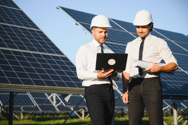 Сосредоточенные инженеры-мужчины в касках с помощью ноутбука и обсуждают проект, стоя рядом с фотоэлектрическими батареями в сельской местности.