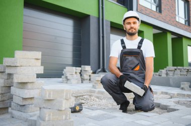 İşçi ustalıkla kaldırım taşları döşeyip çekiç kullanıyor ve eldiven takıyor..