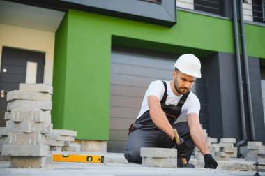 İşçi ustalıkla kaldırım taşları döşeyip çekiç kullanıyor ve eldiven takıyor..