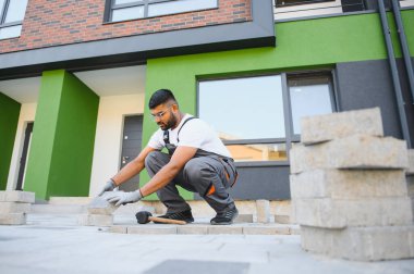 Eldivenli Hintli usta katman katman taş döşüyordu. Profesyonel kaldırım işçisi tarafından kaldırım taşı döşenmiş. Kum temeli üzerine ev avlusunda gri beton döşeme döşeme döşeme döşeme.