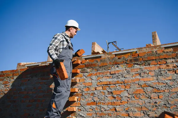 Kırmızı tuğlalı ev inşaatında üniformalı ve kasklı bir erkek inşaat işçisi..