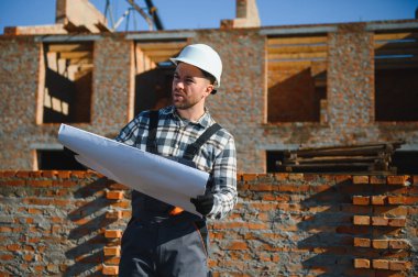 Tuğladan evler inşa eden bir mimar ya da mühendis inşaat planını elinde tutuyor ve işlerin nasıl yürüdüğünü kontrol ediyor..