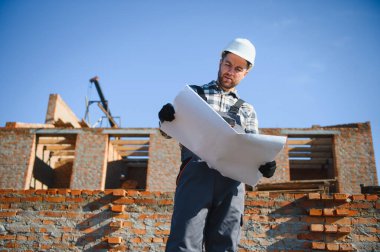 Tuğladan evler inşa eden bir mimar ya da mühendis inşaat planını elinde tutuyor ve işlerin nasıl yürüdüğünü kontrol ediyor..