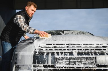 Adam araba yıkama servisini köpük ve süngerle yıkıyor. Araba yıkama istasyonu. Çalışanlar bir aracı profesyonel olarak temizler.