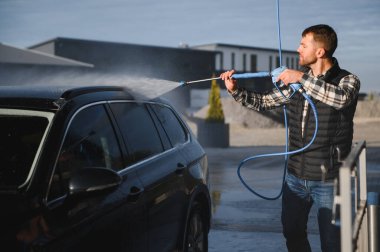 Bir adam arabasını tazyikli suyla yıkama makinesinde yıkıyor..