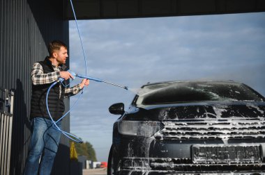 Bir adam arabasını tazyikli suyla yıkama makinesinde yıkıyor..