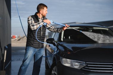 Bir adam arabasını tazyikli suyla yıkama makinesinde yıkıyor..