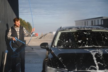 Bir adam arabasını tazyikli suyla yıkama makinesinde yıkıyor..