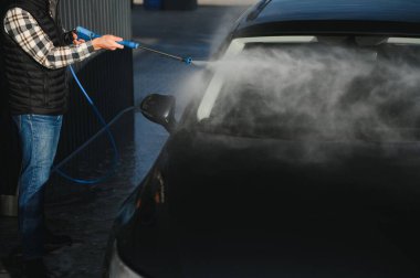 Bir adam arabasını tazyikli suyla yıkama makinesinde yıkıyor..