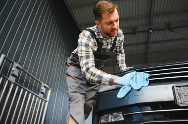 Sakallı oto yıkama işçisi, koruyucu kıyafetler giyiyor üniformalar giyiyor, modern arabayı mikrofiber bezle temizliyor..