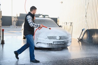 Bir adam arabasını tazyikli suyla yıkama makinesinde yıkıyor..