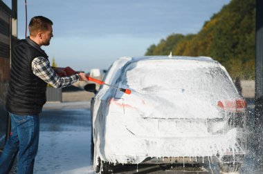 Bir adam arabayı yıkarken köpüğü yıkar. Bir araç araba yıkama sırasında sabunlu köpüğe bulanır, lastikleri, tekerlekleri, kaputu ve arabanın tüm dış kısmını kaplar. self-servis araba yıkama