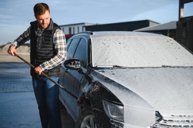 Bir adam arabayı yıkarken köpüğü yıkar. Bir araç araba yıkama sırasında sabunlu köpüğe bulanır, lastikleri, tekerlekleri, kaputu ve arabanın tüm dış kısmını kaplar. self-servis araba yıkama