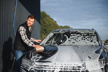 Adam araba yıkama servisini köpük ve süngerle yıkıyor. Araba yıkama istasyonu. Çalışanlar bir aracı profesyonel olarak temizler.