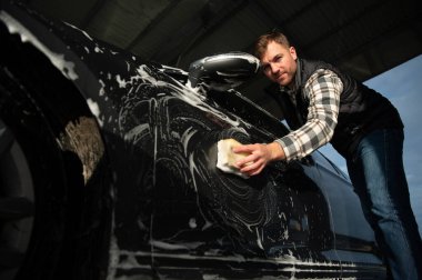Adam araba yıkama servisini köpük ve süngerle yıkıyor. Araba yıkama istasyonu. Çalışanlar bir aracı profesyonel olarak temizler.