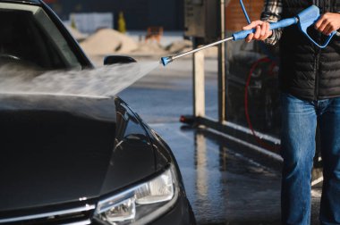 Bir adam arabasını tazyikli suyla yıkama makinesinde yıkıyor..