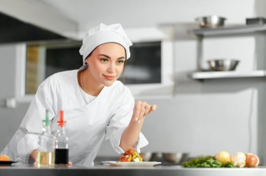 Genç, güzel, gülümseyen kadın şef mutfakta beyaz takım elbise giyiyor. Bulanık arkaplan.