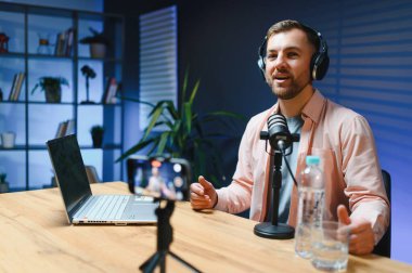 Adam podcaster etkili blogcu gülümseyerek stüdyoda kulaklık, dizüstü bilgisayar ve kulaklık kullanarak canlı yayın yapıyor. Erkek radyo sunucusu podcast veya röportaj yapıyor