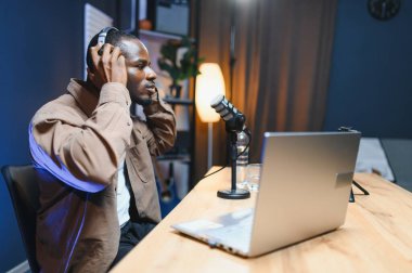 Genç Afro-Amerikalı erkek video blogcu kulaklıklarda etkili. Evdeki akıllı telefondan video blogu kaydediyor. Milenyum melez erkek blogcu konuşması sosyal medya için cep telefonundan canlı yayın..