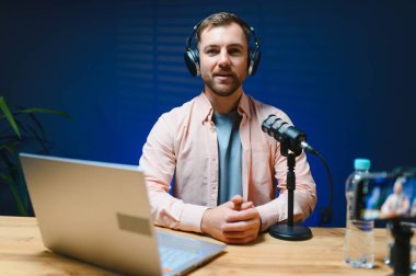 Adam podcaster etkili blogcu gülümseyerek stüdyoda kulaklık kullanarak canlı yayın yapıyor. Erkek radyo sunucusu podcast veya röportaj yapıyor.