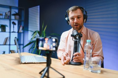 Adam podcaster etkili blogcu gülümseyerek stüdyoda kulaklık kullanarak canlı yayın yapıyor. Erkek radyo sunucusu podcast veya röportaj yapıyor.