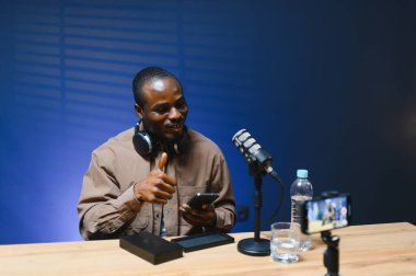 Siyah vlogger stüdyoda akıllı telefondan podcast çekiyor. Afro-amerikan etkileyicisi telefon ekranına bakıyor ve iyi haberleri seyircilere ve abonelere veriyor. Neşeli blogcu
