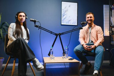 Kulaklıklı kadın podcaster mikrofon kullanarak, gülümseyerek, kayıt yaparak ve stüdyoda ses yayını yaparak erkek konukla röportaj yapıyor. İçerik oluşturucu, günlükler.