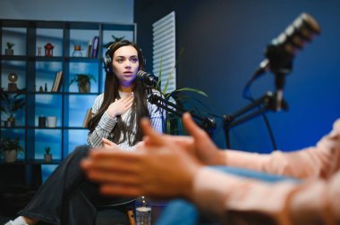 Kulaklıklı kadın podcaster mikrofon kullanarak, gülümseyerek, kayıt yaparak ve stüdyoda ses yayını yaparak erkek konukla röportaj yapıyor. İçerik oluşturucu, günlükler.