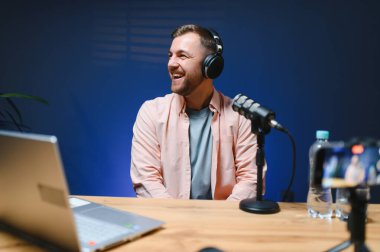 Adam podcaster etkili blogcu gülümseyerek stüdyoda kulaklık, dizüstü bilgisayar ve kulaklık kullanarak canlı yayın yapıyor. Erkek radyo sunucusu podcast veya röportaj yapıyor