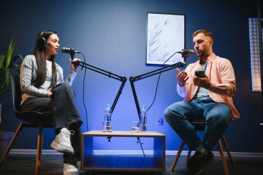 Kulaklıklı kadın podcaster mikrofon kullanarak, gülümseyerek, kayıt yaparak ve stüdyoda ses yayını yaparak erkek konukla röportaj yapıyor. İçerik oluşturucu, günlükler.