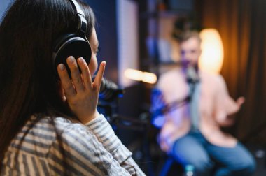 Kulaklıklı kadın podcaster mikrofon kullanarak, gülümseyerek, kayıt yaparak ve stüdyoda ses yayını yaparak erkek konukla röportaj yapıyor. İçerik oluşturucu, günlükler.