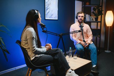 Kulaklıklı kadın podcaster mikrofon kullanarak, gülümseyerek, kayıt yaparak ve stüdyoda ses yayını yaparak erkek konukla röportaj yapıyor. İçerik oluşturucu, günlükler.