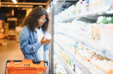 Market alışverişi yapan, alışveriş yapan, yiyecek seçen ve alışveriş yapan mutlu siyah kadın. Modern süpermarketin raflarında market arabasıyla ayakta duruyor..